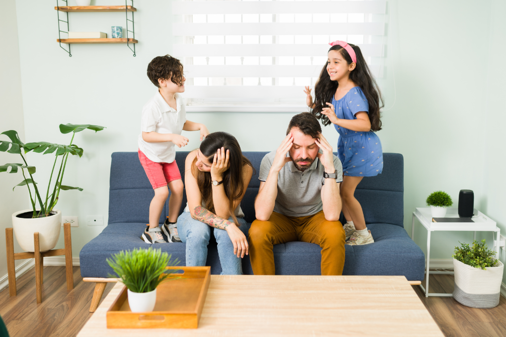 Stressed young parents feeling tired while their children jump on a couch around them