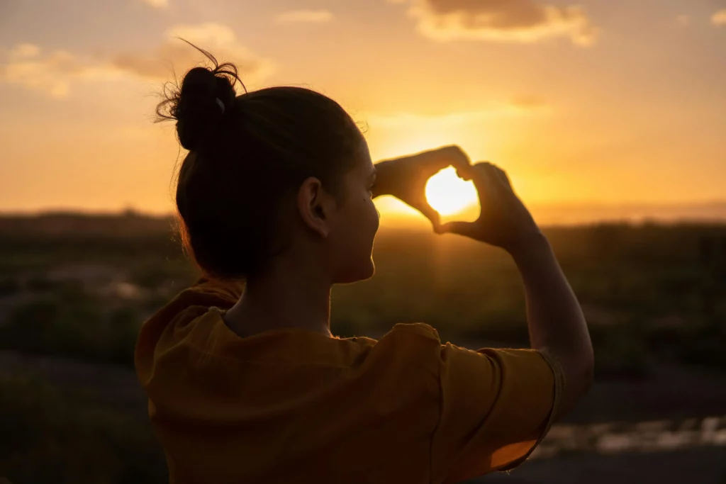 Woman making a heart shape with her hands in front of a sunset